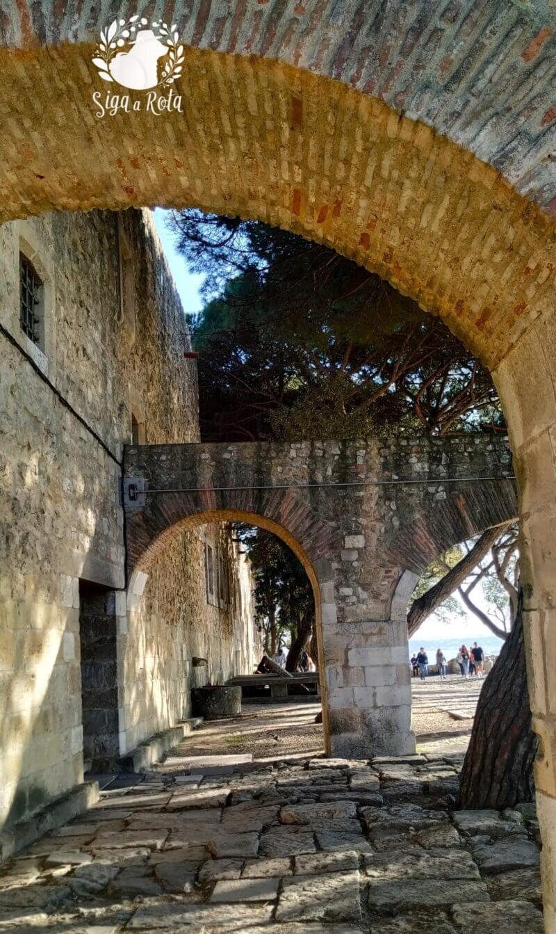 Área de jardins e muralhas no Castelo de São Jorge, com árvores e vista para Lisboa.