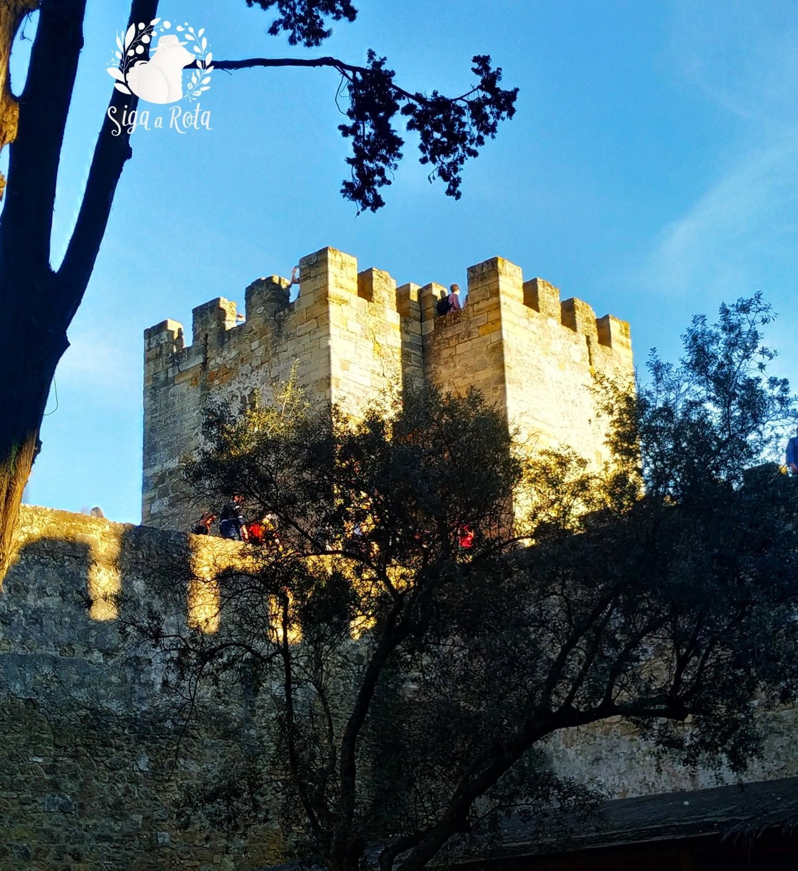 Muralhas do Castelo de São Jorge iluminadas pelo pôr do sol, com vista panorâmica da cidade.