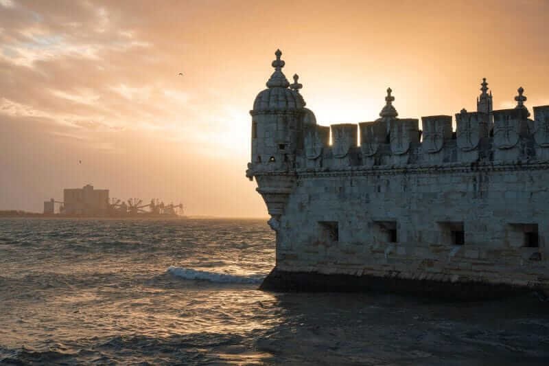 "Torre de Belém iluminada pelo pôr do sol, com tons alaranjados refletidos nas águas do Rio Tejo.