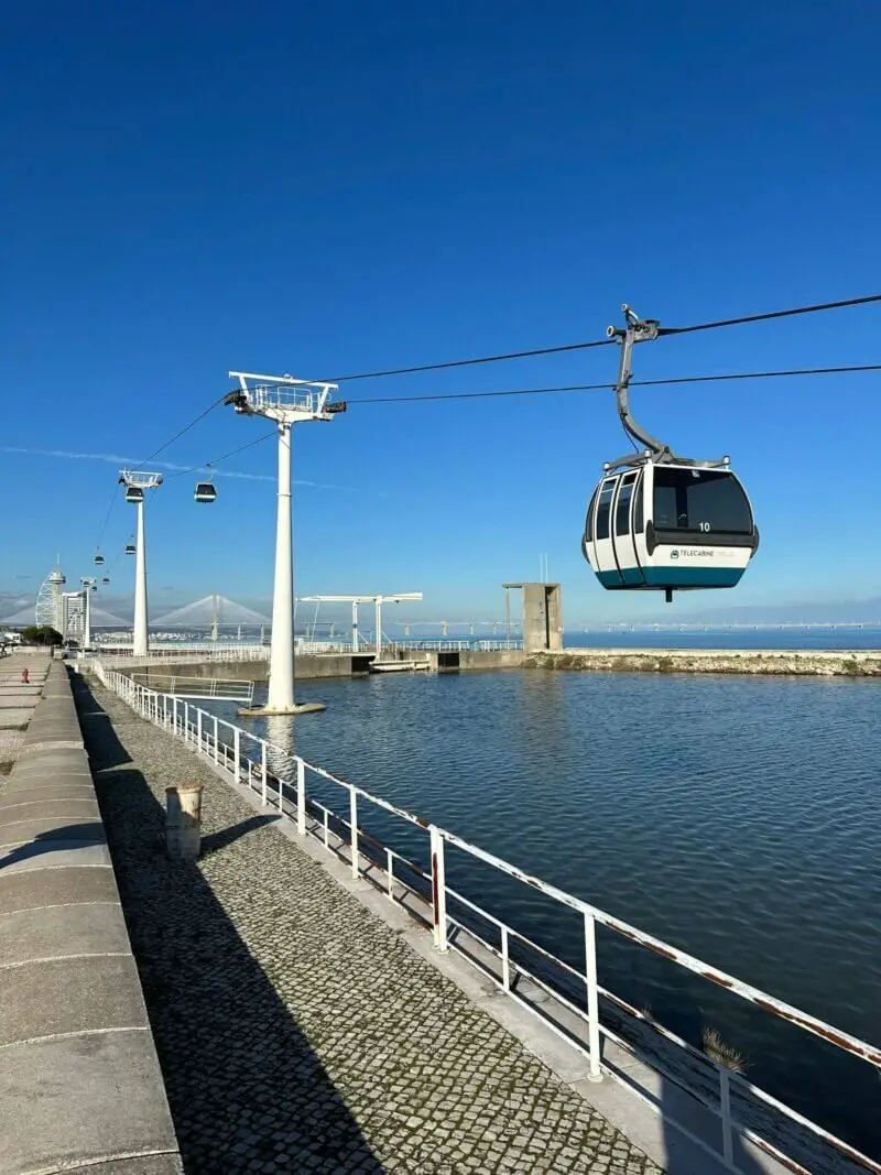 Vista do teleférico em movimento sobre o Parque das Nações, com destaque para as modernas instalações e o rio Tejo.