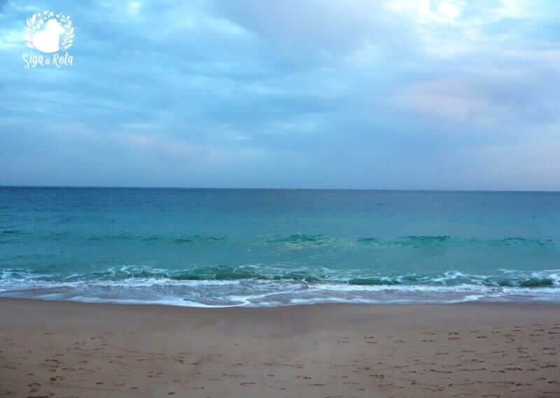 Vista panorâmica da Praia do Ouro em Sesimbra, com a Fortaleza de Santiago ao fundo, destacando sua posição estratégica à beira-mar.