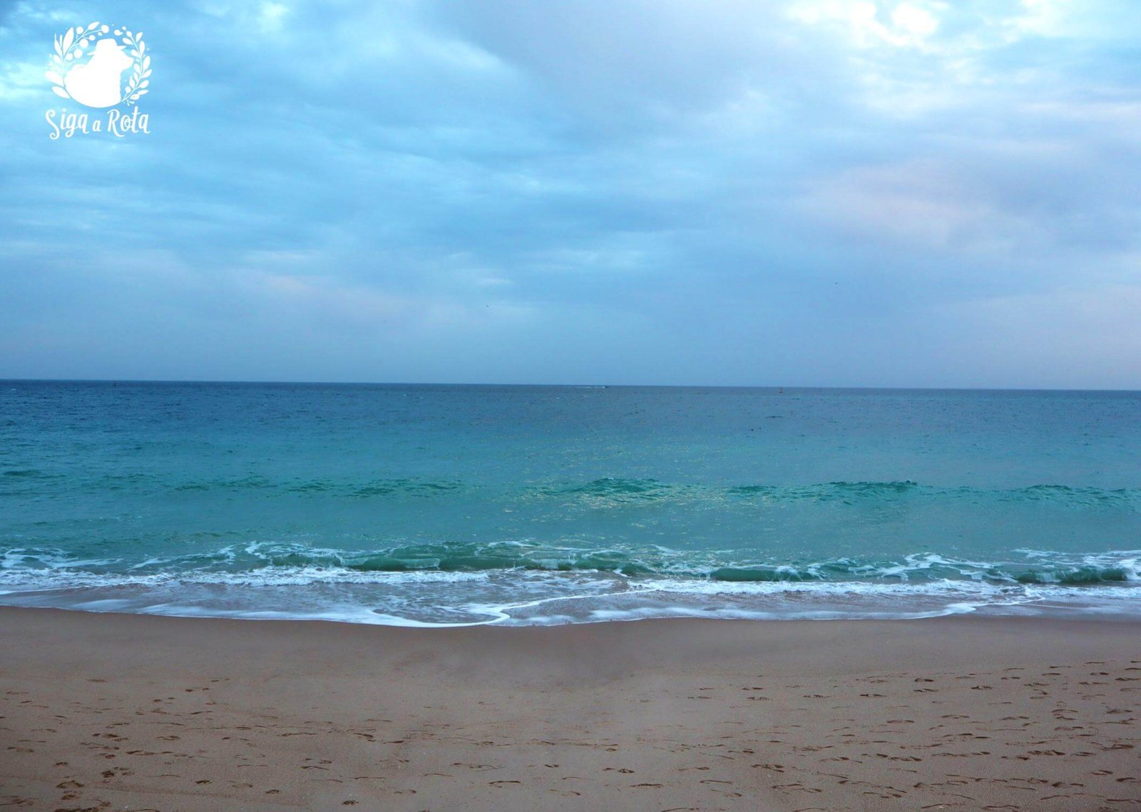 Vista panorâmica da Praia do Ouro em Sesimbra, com a Fortaleza de Santiago ao fundo, destacando sua posição estratégica à beira-mar.